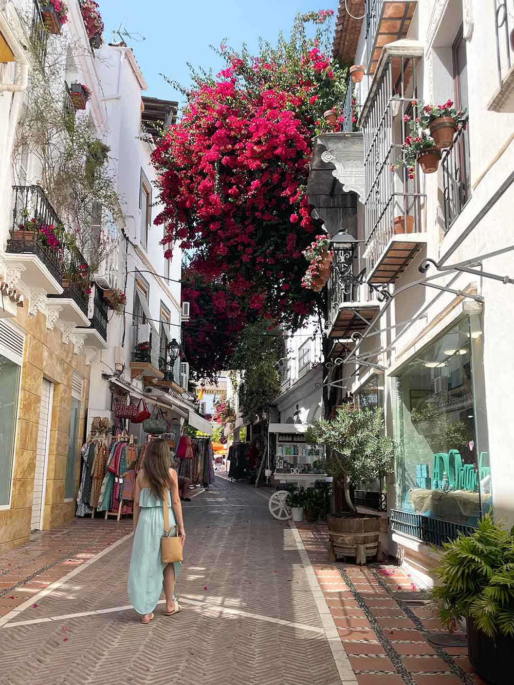 A woman in a light blue dress a walks down a quaint cobblestone street lined with white-walled shops and pink bougainvillea cascading from an upper balcony in Marbella Old Town