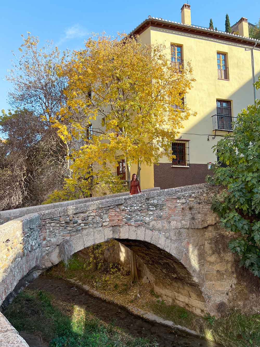 Stone bridge with an arch over a small stream flanked by yellow autumn trees and greenery, with Cristina standing on the bridge.
