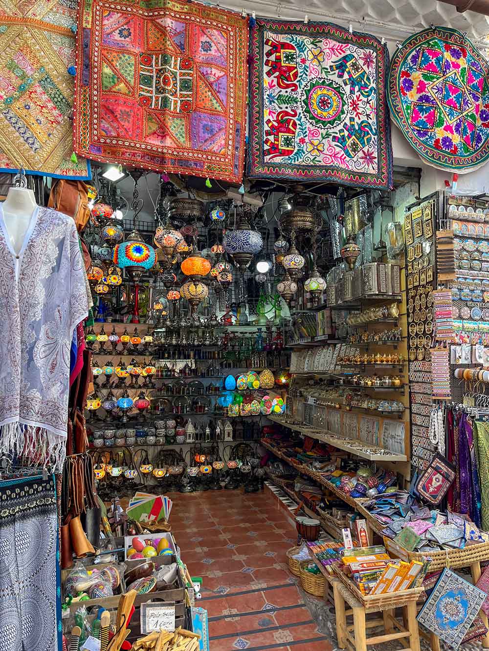 Cosy ethnic shop interior packed with an eclectic mix of items, featuring colorful embroidered tapestries, intricate glass lanterns, a variety of ceramics, and cultural souvenirs.