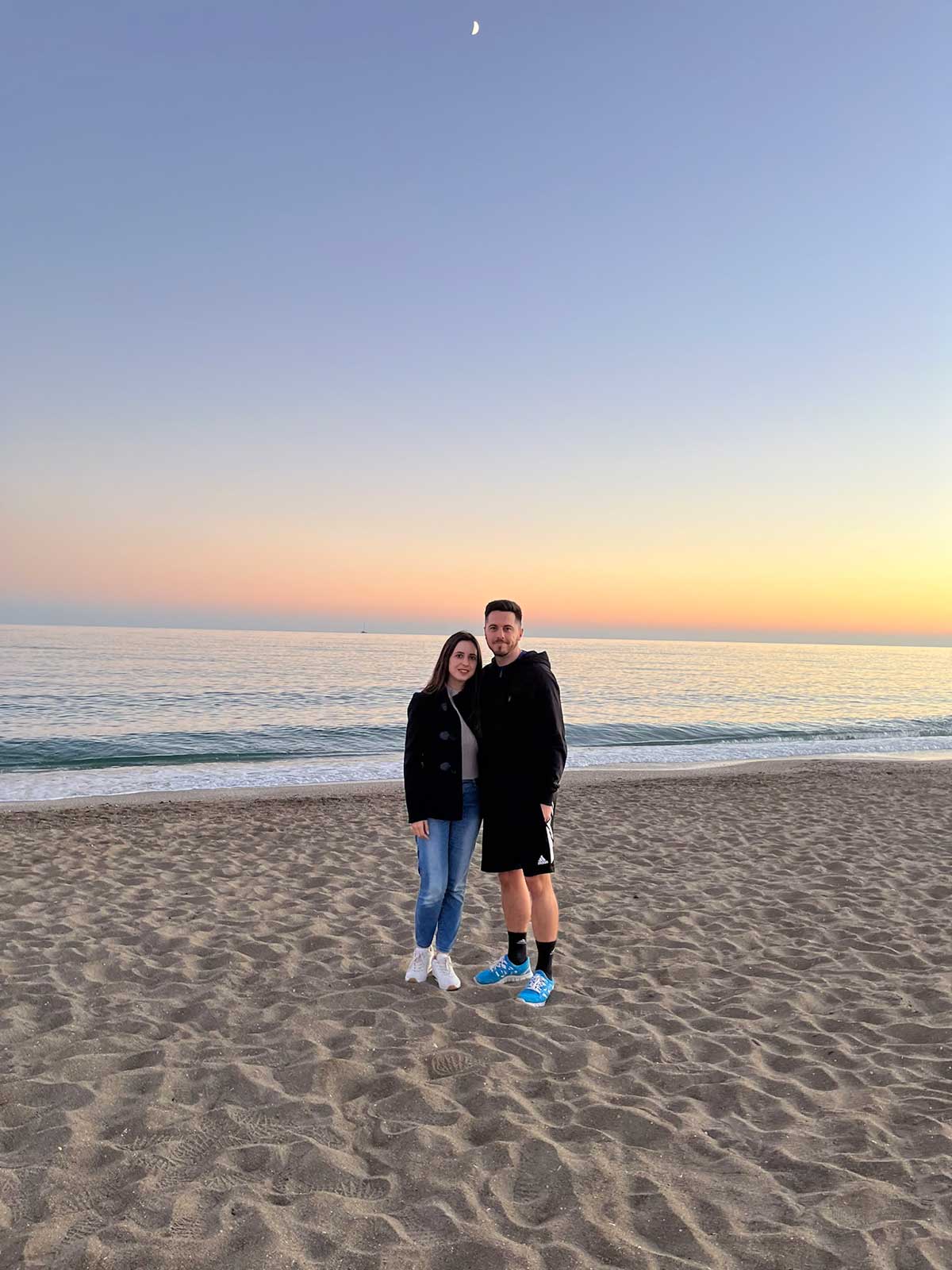 A man and a woman standing on a sandy beach in Mijas with the ocean in the background during sunset, with a clear sky and a crescent moon visible above.
