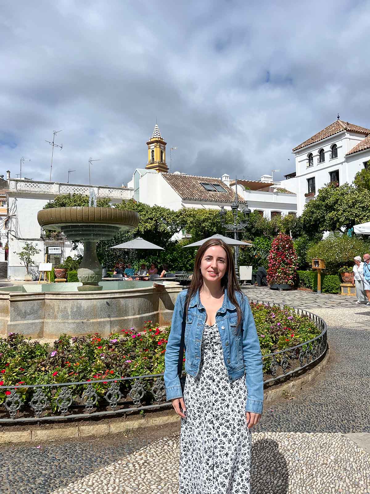 Cristina wearing a black and white dress with a denim jacket posing in front of the fountain in Plaza de las Flores, Estepona.