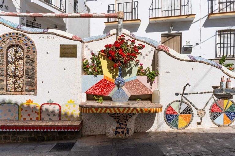 Colorful mosaic-tiled public fountain and benches on a sunny street in Moclinejo, Spain. The fountain features a geometric star design with red flowers in the center, surrounded by bright tiles in blue, yellow, red, and pink. A mosaic bicycle design decorates the wall beside it, and a plaque with an inscription is mounted above the bench on the left.