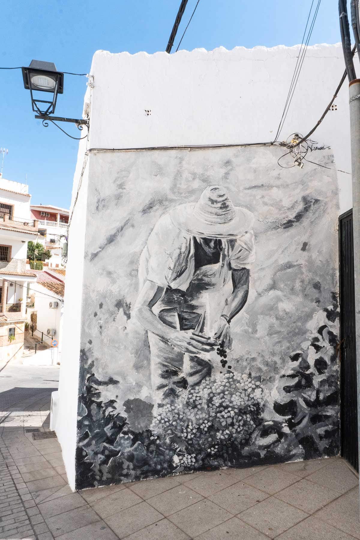 Black and white mural painted on the side of a white building, depicting a person wearing a hat and bending down to pick grapes or plants. The mural is located on a street corner in a small town with white houses and tiled roofs under a clear blue sky.